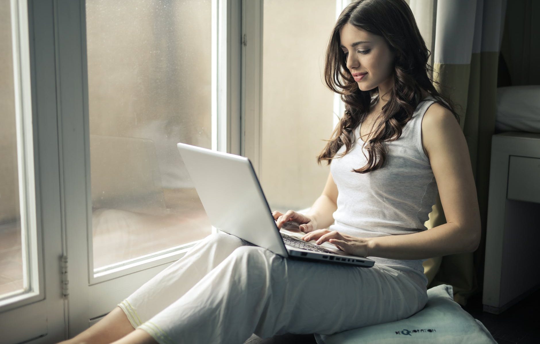 Frau sitzt am Fenster auf einem Kissen und surft mit einem Notebook im Web, sieht sich Webseiten an. Sie macht sich Gedanken über die Zukunft des Webs.