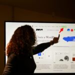 A person with curly hair is silhouetted against a large computer screen, pointing at a section of a webpage. The screen displays a website in Spanish, featuring the WordPress logo and navigation menu, with a red arrow pointing to the “Colabora” menu item.